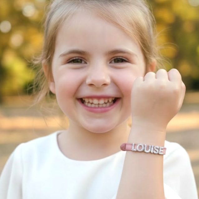 Avis Client Bijou Enfant - Rêve Étoilé Avis client Rêve Étoilé avec une petite fille souriante portant un bracelet personnalisé en cuir rose au prénom de Louise
